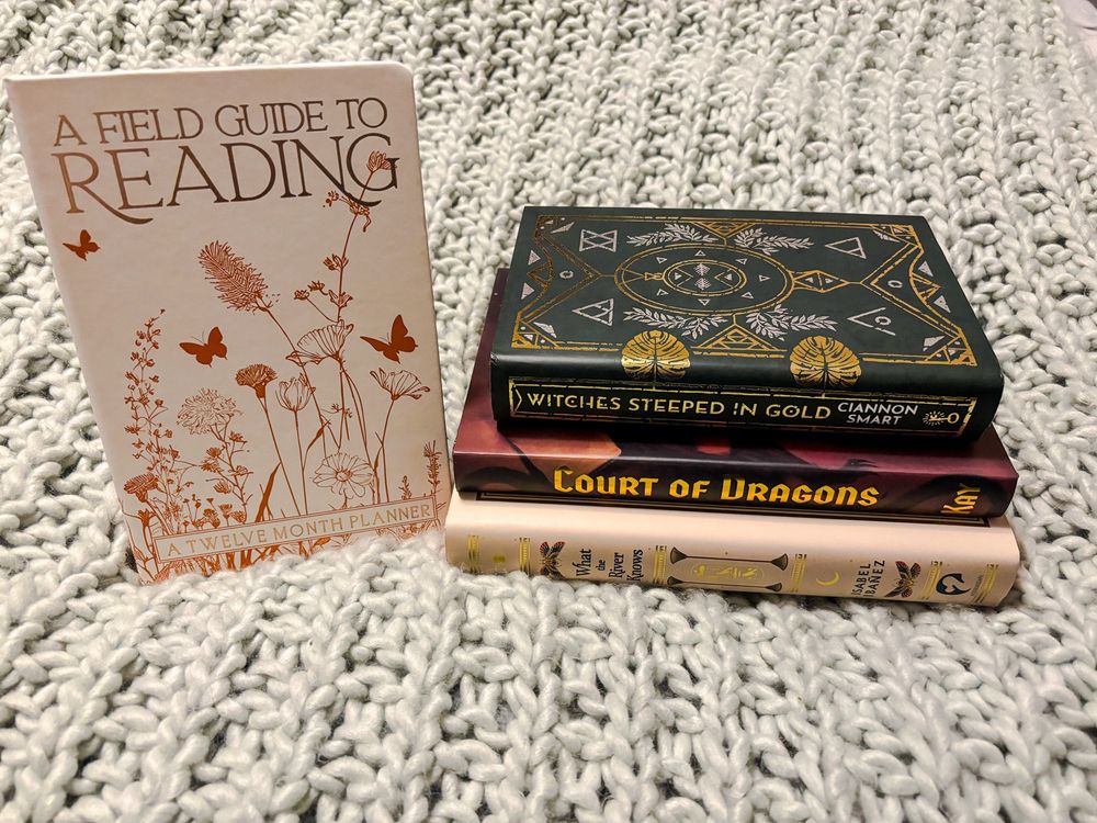 A stack of 3 books: What the River Knows by Isabel Ibanez, Court of Dragons by Kay Ivey, Witches Steeped in Gold by Ciannon Smart. Standing next to the stack is a twelve month reading planner titled “A Field Guide to Reading” it is decorated in a gold floral pattern.