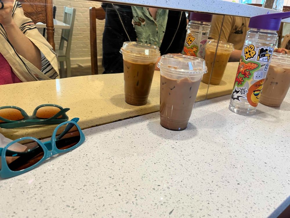 candid photo of a cafe bar with 3 people in the reflection of the wall, their faces aren't visible but their teas/coffees and water bottles are on the surface of the counter itself