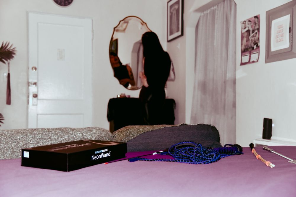 A photo of the view from the bed. In the foreground, a neon wand and various impact toys are in focus. In the background, I'm fixing my makeup in the vanity mirror. 