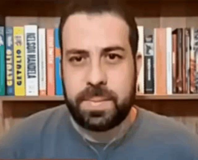 a man with a beard is looking at the camera in front of a bookshelf filled with books .