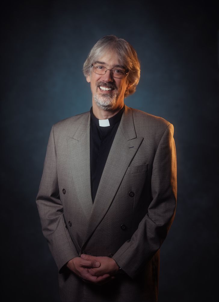 A portrait of James—middle-aged white guy with beard and glasses—wearing a clerical collar and double-breasted suit jacket. 