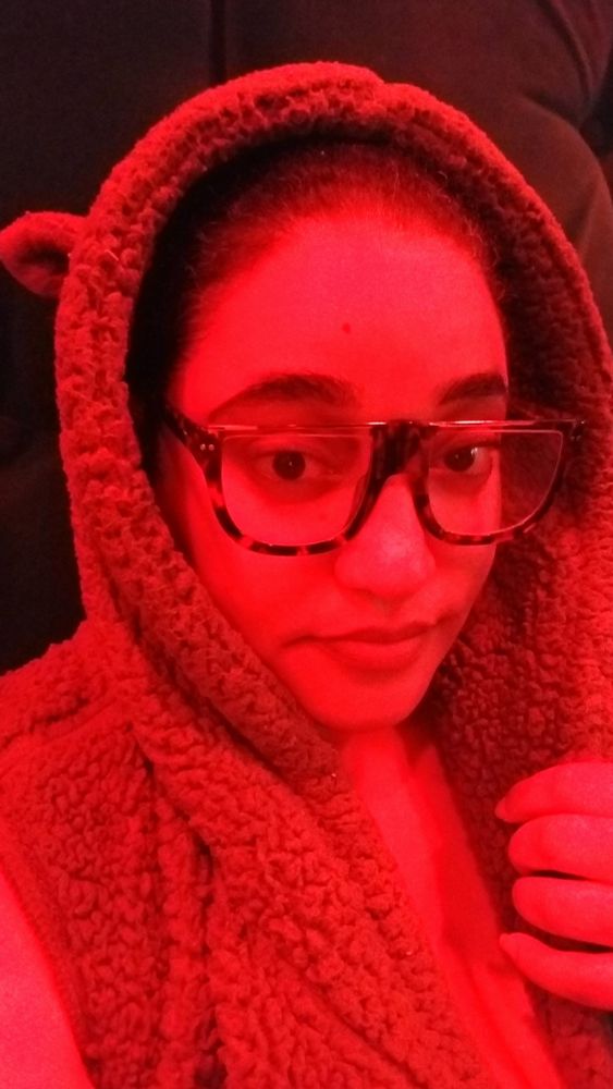 A picture of Jtwiced cast under a red light sitting on a plush backed bench wearing a plush teddy bear robe with ears. She has on brown tortoise shell glasses, rounded on the bottom with gold bars going across the top of each eyeglass.