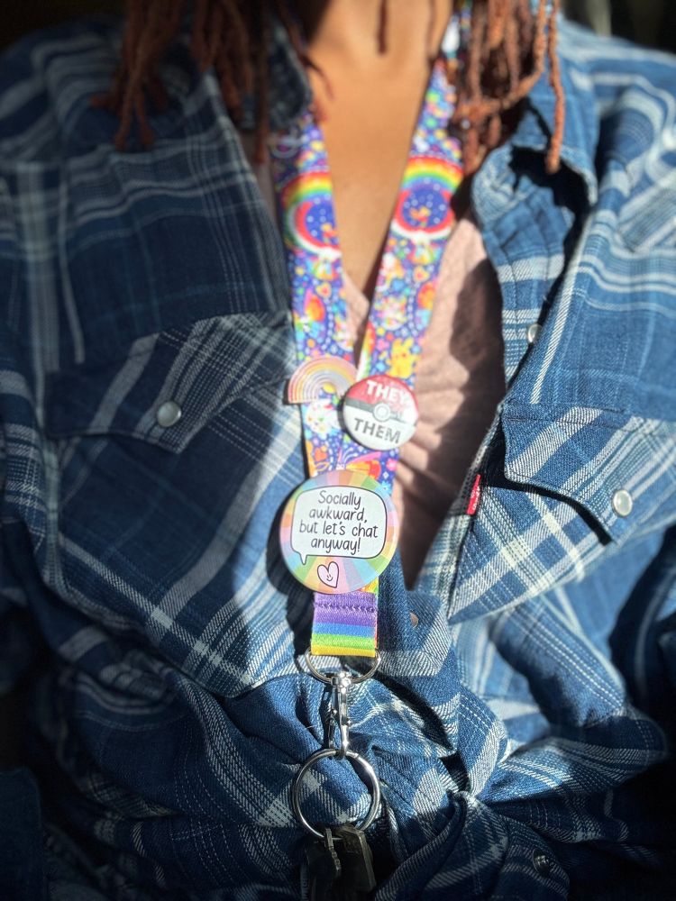 Picture is of a key lanyard with rainbows and a blue background. The button on the bottom says socially awkward, but let’s chat anyway there is another smaller button with a Poké ball and the pronoun gave them and then there is a small rainbow button.