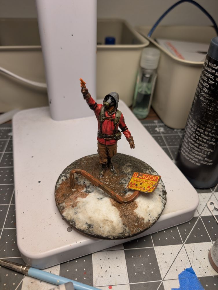 A small figure with a gas mask and a red jacket on a muddy, snowy base with a radiation warning sign tipped over nearby