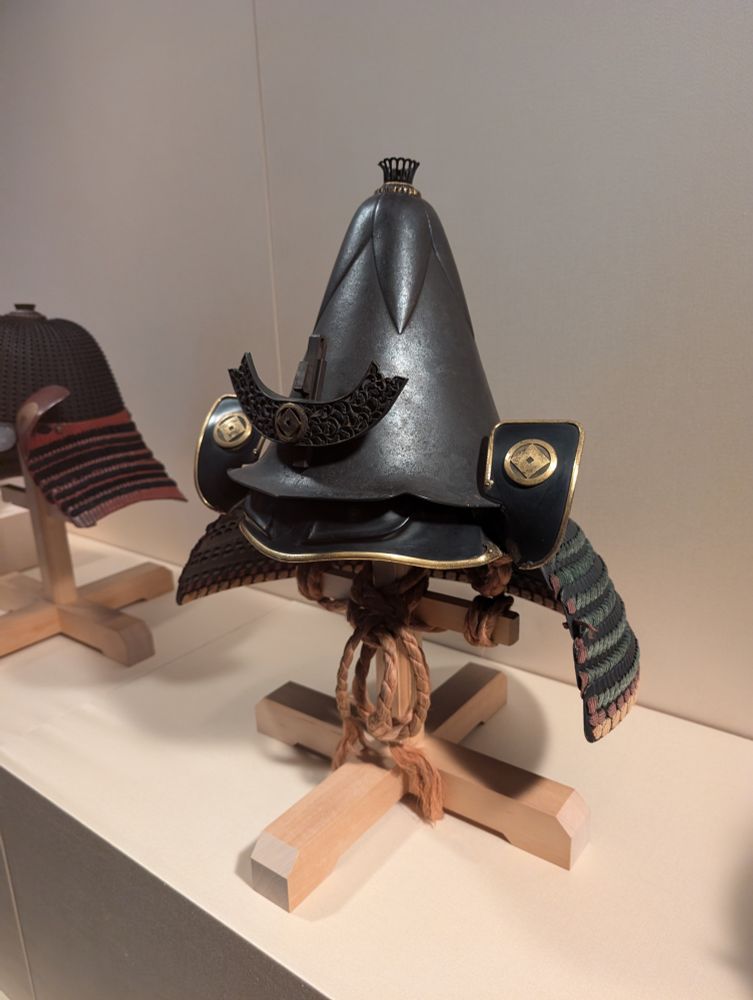 A shot of an eggplant-shaped samurai helmet sitting on a wooden stand in a museum display case. It's made of darkened metal and has gold trim and accents.