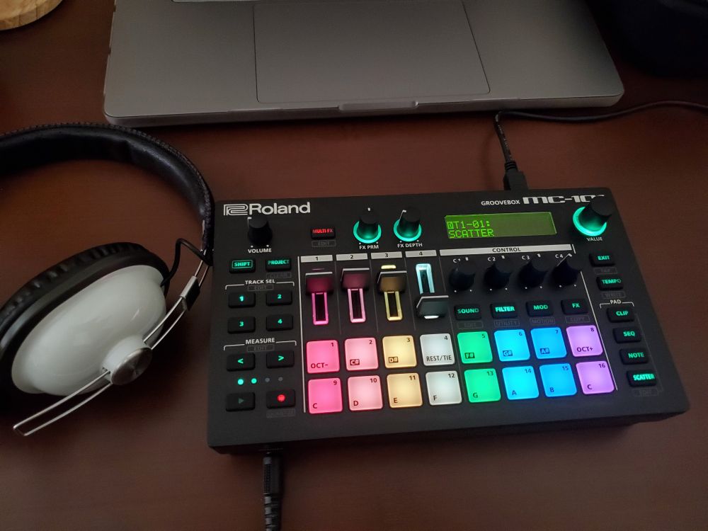 A Roland MC-101 groovebox, some really old headphones, and a bit of a Macbook on a cheap desk.