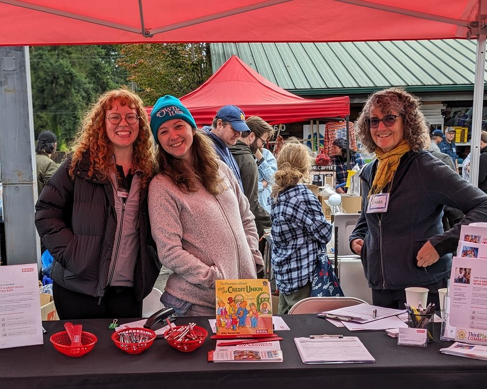 Two folks from BECU and another volunteer smile under the BECU tent. Folks exploring the Shoreline Tool Library Tool Sale walk behind them.
