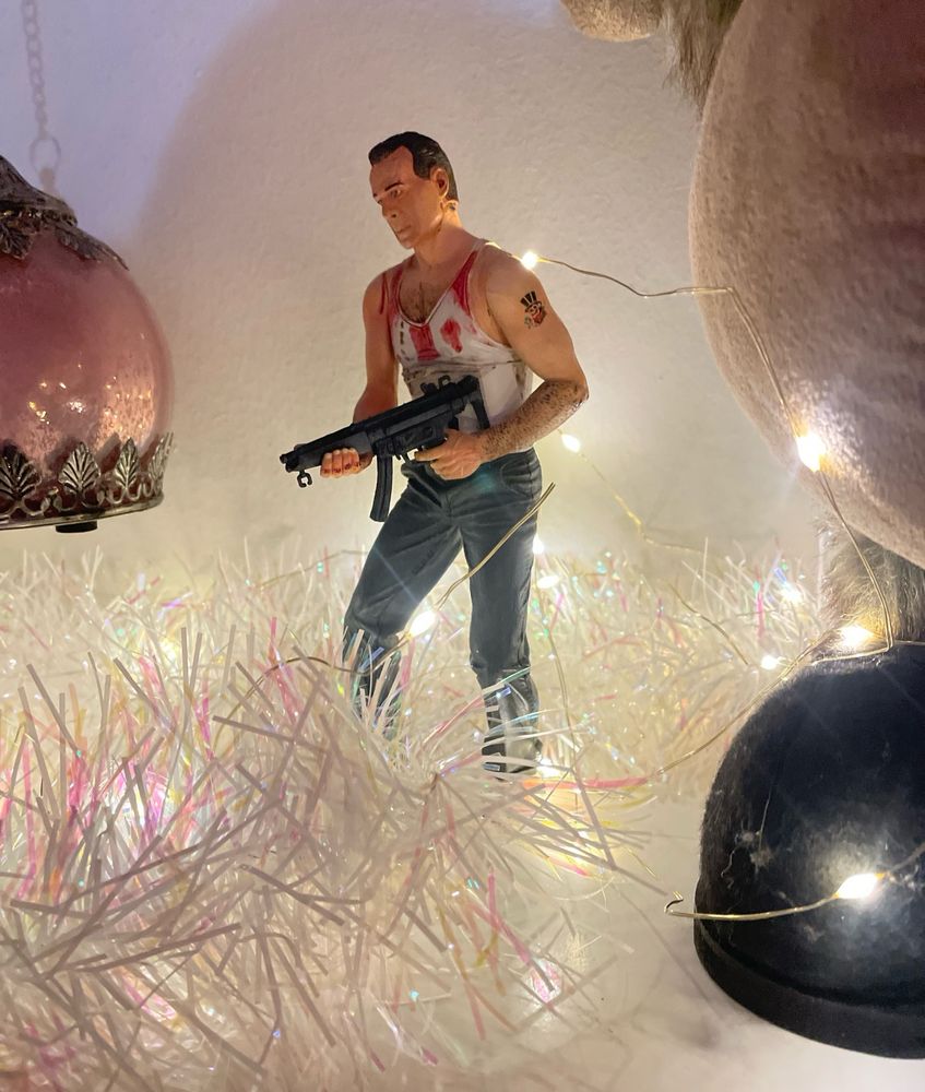 John McClane figurine, from Die Hard movies. White undershirt, bloodstained, holding machine gun, standing on a white dresser amonst white Christmas lights and tinsel.