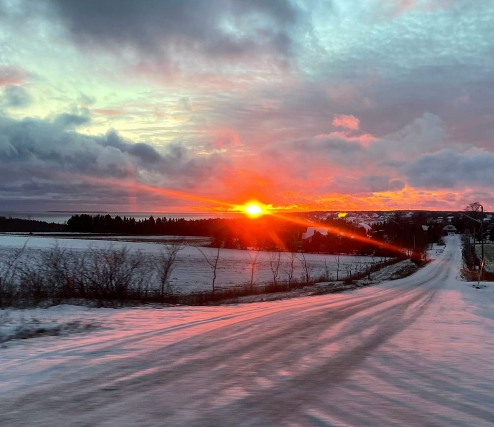 The sun setting on the ocean, behind a long road covered in a light dusting of snow.