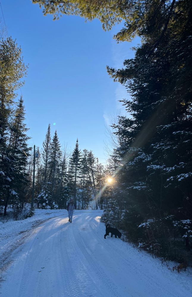My young daughter adjusting her kitbag over the cast on her arm, her black dog floofing around the white snowy lane in front of her. The sun is setting behind her over the snow covered trees.
