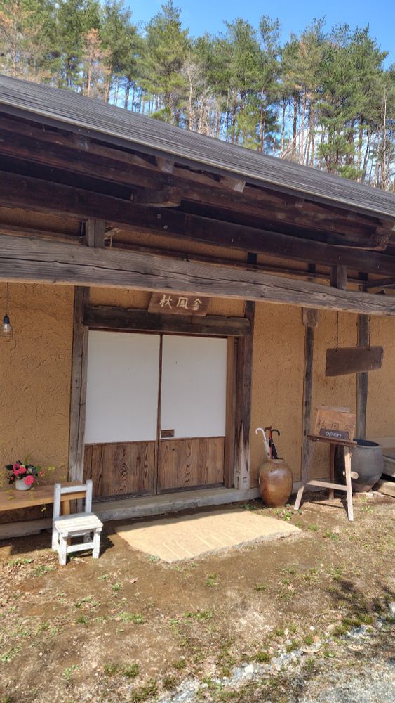 カフェ「秋風舎」外観。庵風の平屋。土壁の真ん中に引き戸、上に気の看板が掲げてある。向かって左には小さな白い椅子と生け花、向かって右には大きな陶器の壺（傘立て）とイーゼル。イーゼルには「秋風舎 OPEN」と掲げられている