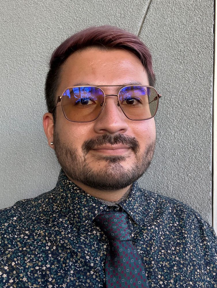 Selfie of me smiling into the camera. My hair is now a fuchsia color. My glasses are sight tinted brown. I’m wearing a green shirt with small flowers and a patterned tie