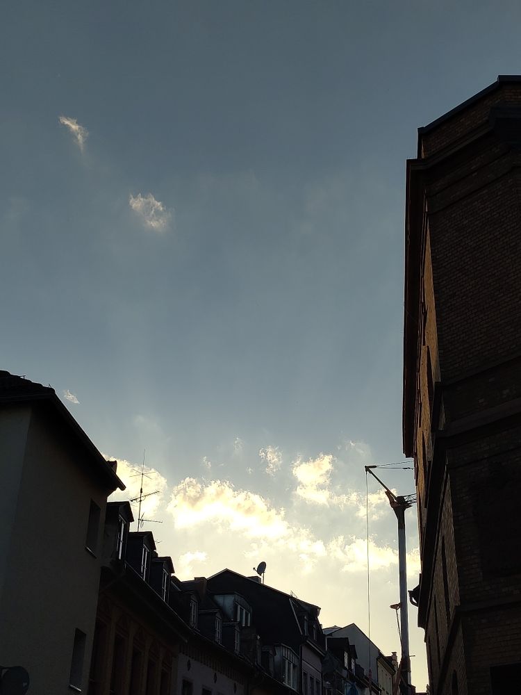 Photo of a blue sky with a few golden-white clouds near the horizon. Below and on the right are buildings.