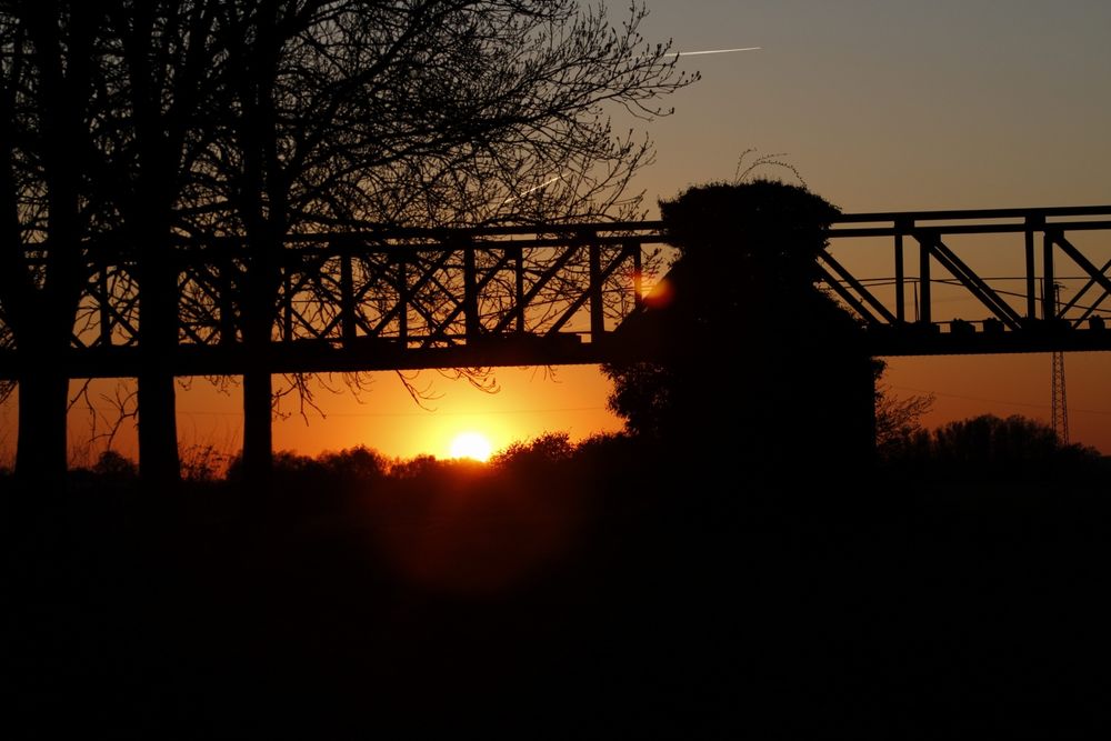Sonnenuntergang an einer alten Eisenbahnbrücke. Links im Bild 3 Bäume, Die Eisenbahnstrecke teilt das Bild in der waagerechten. In der Mitte sieht man einen Stützpfeiler für die Brücke. Die Sonne ist bereits zur Hälfte am Horizont verschwunden.