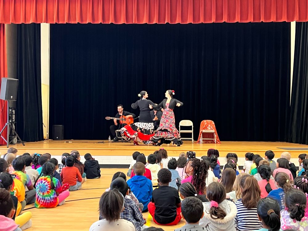Two flamenco dancers in long flowered dresses link arms while dancing. Their troopmate plays guitar behind them. Elementary school children sit on the floor watching.