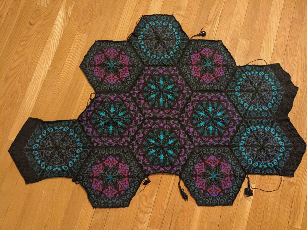 A knitted blanket on a wood floor. The blanket is partially complete, made of 12 (so far) tiling hexagons with a black background and abstract/floral patterns in purples and teals. Three of the outer hexes have thick black edging on two sides, but all the other edges are unfinished and awaiting more hexes. Unfinished ends trail off of many of the edges.