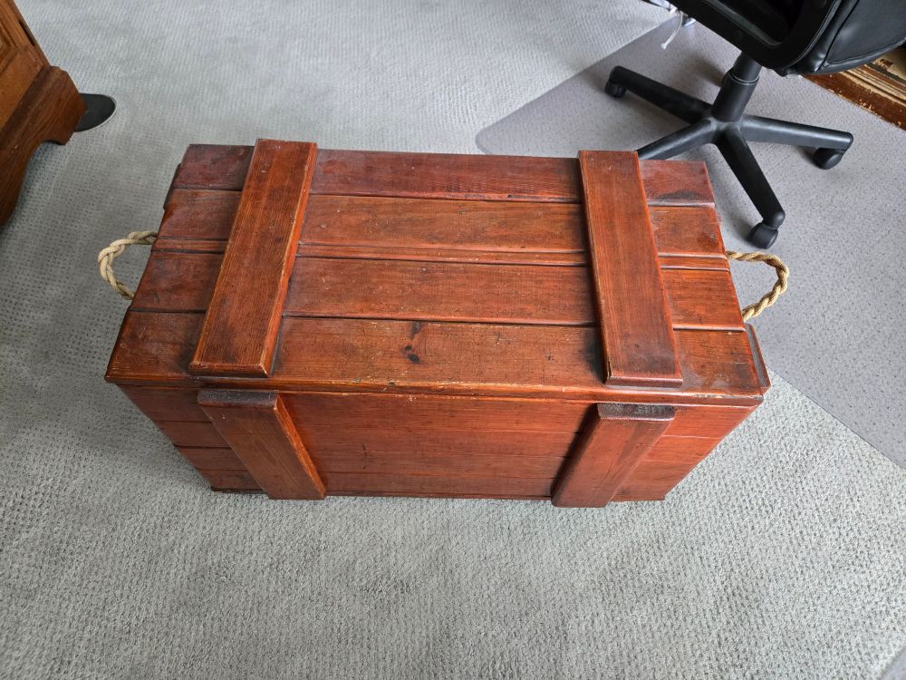 A dark stained crate-like wooden chest with rope handles sitting on grey carpeting with a computer chair on a mat in the upper right corner. 4 long boards make up the top and are secured to each other with 2 narrow boards along each side.