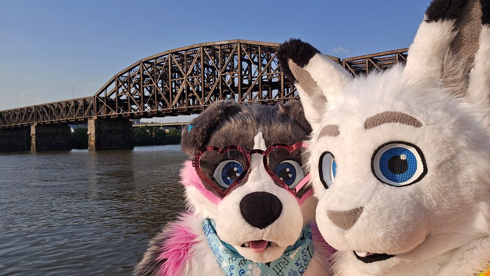 2 fursuiters standing in front of a steel train trestle bridge over the Allegheny River. The one on the left is a grey, white, pink, and blue dog wearing a blue bandana and pink reflective heart shaped glasses. The one on the right is a white hare with black ear tips and blue eyes. A safety vest can be barely seen in the bottom right corner.