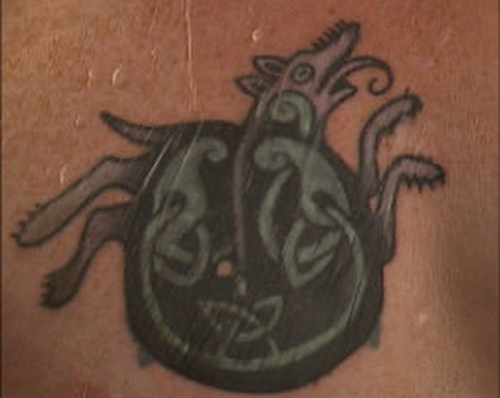 graham platners coverup tattoo. a black splotch with green designs that seem meant to look like celtic knotwork. sticking out from behind the splotch is a dog or wolf-like figure. the lower corners of the original totenkopf can be seen poking out from behind.