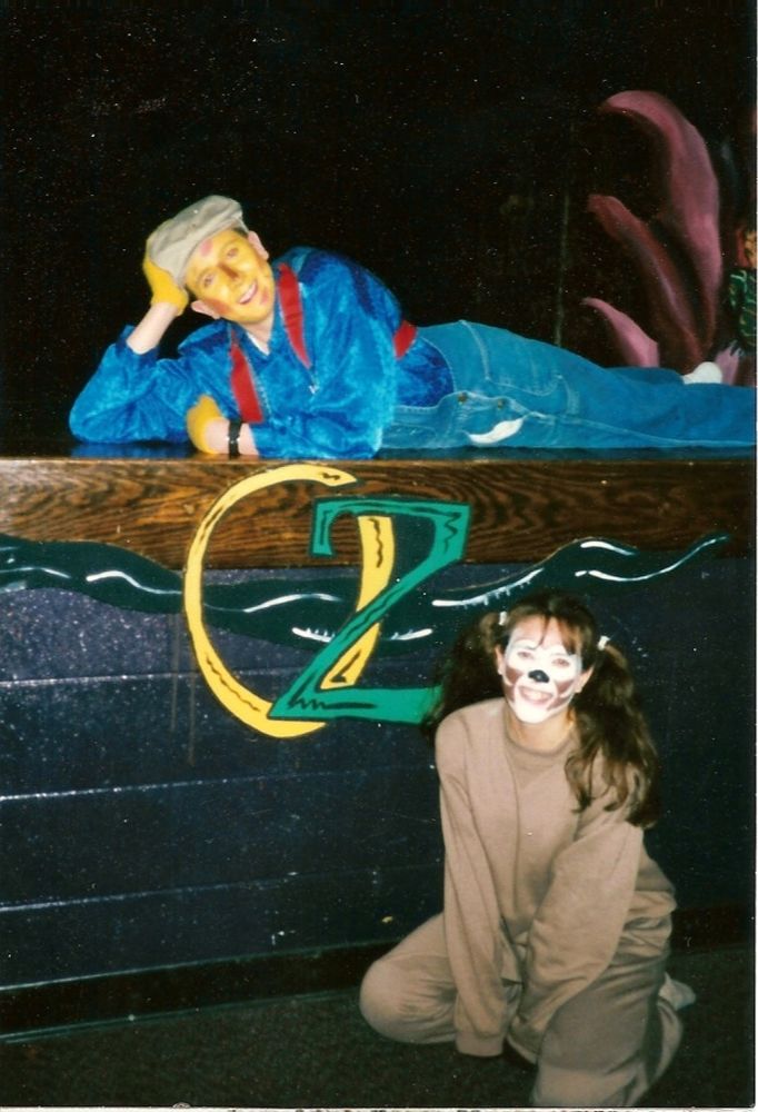 teenage kelcy dressed as the scarecrow laying on the stage above a yellow and green OZ logo with her head propped up on her hand. her sister crouches below dressed as toto