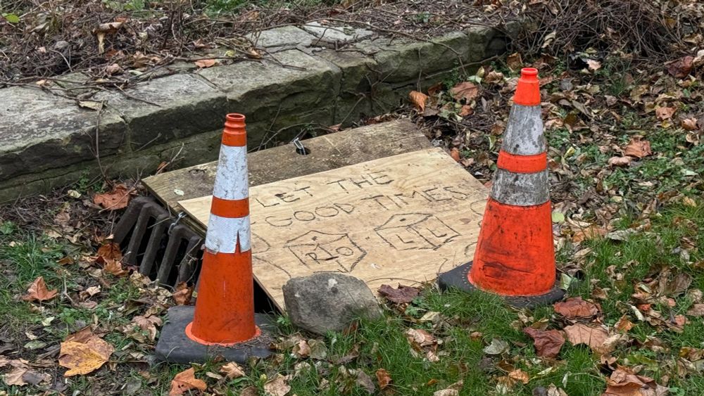 A wooden board blocking a sewer grate has "LET THE GOOD TIMES ROLL" written on it. The "ROLL" is written on a pair of dice.