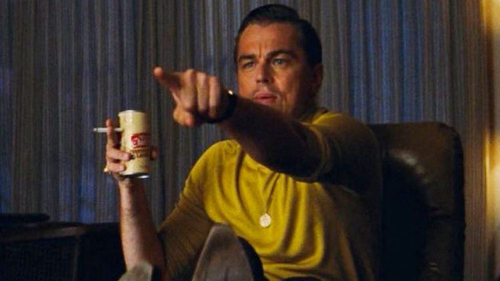 Leonardo DiCaprio sitting on a comfy chair, holding a beer, pointing in recognition at something offscreen