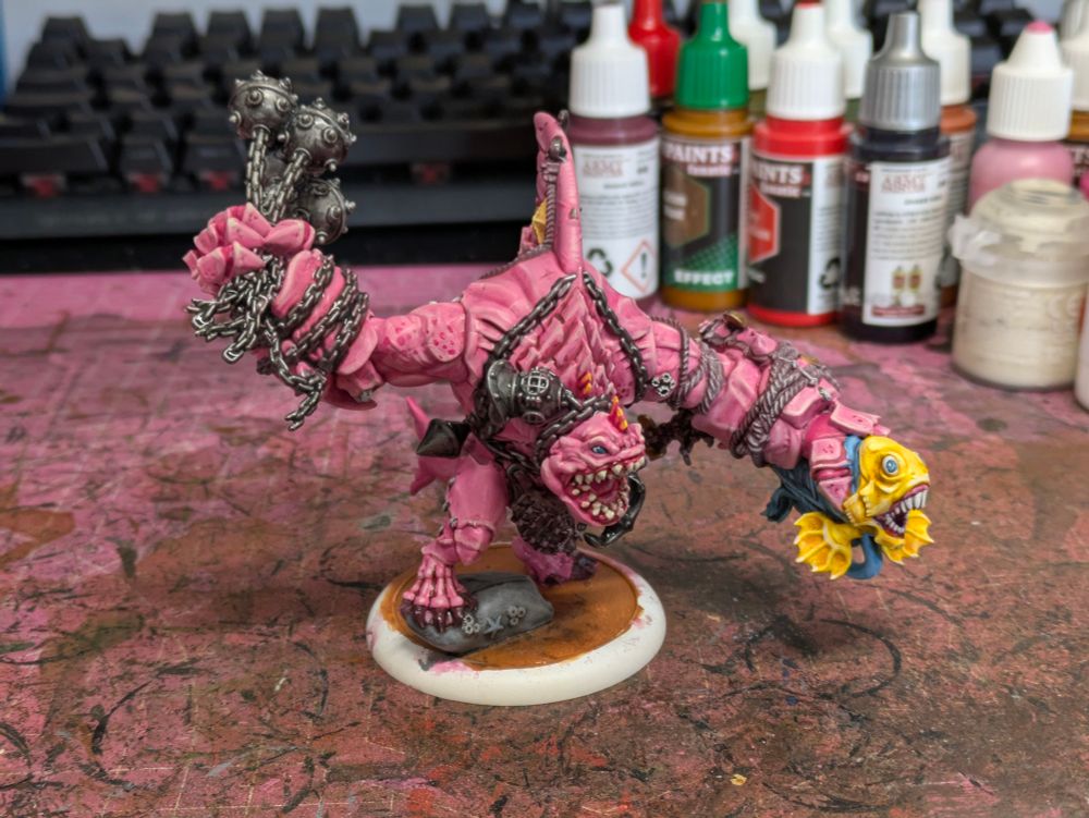 A pink sharkman holding a yellow and blue fish on one hand, and a flail made of sea mines and chains in the other. 