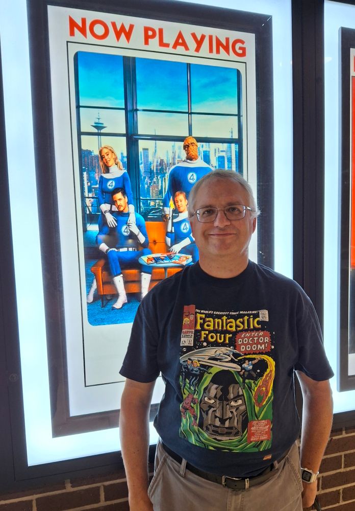 A nerdy, gray-haired, white dude wearing a Fantastic Four t-shirt in front of the "Fantastic Four: First Steps" movie poster.