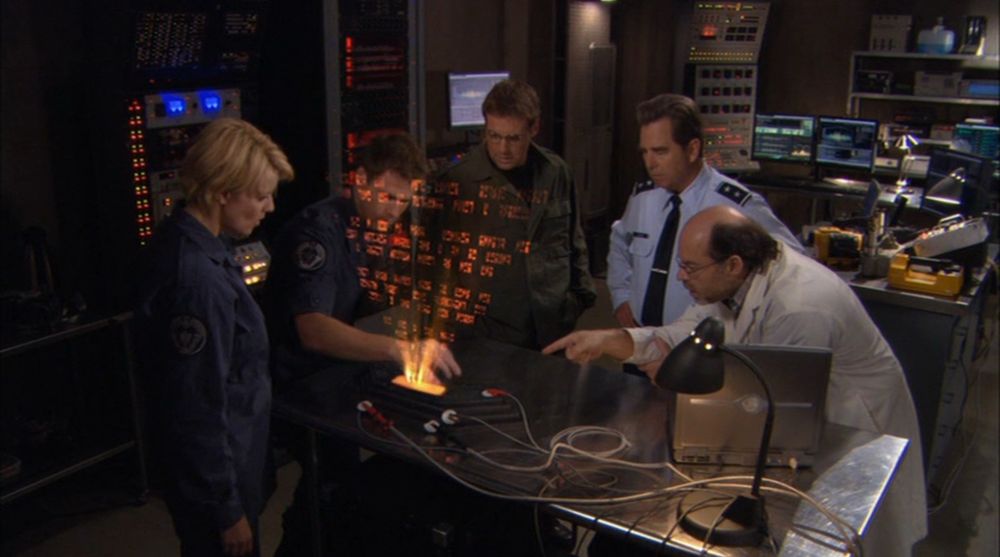 Sam, Cam, Daniel, Landry, and Dr. Lee are all standing around an Ancient keyboard with an orange holographic display being projected from it. They’re in Dr. Lee’s office so there are lots of computers, machinery, and scientific equipment around the room. The keyboard is on a metal table with electrodes attached to it and a laptop and lamp also on the table. Cam is pressing a key on the keyboard and Dr. Lee is pointing at what he’s doing. You can’t tell from the photo but Sam and Cam are out of phase! Arezou and I spend several minutes of our podcast discussing how you would go to the bathroom if you were out of phase…. 😂 