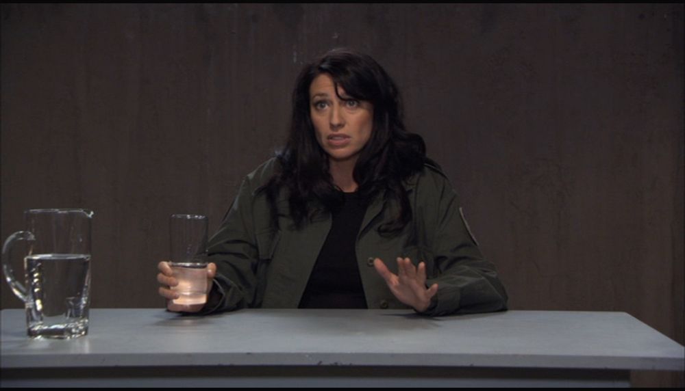 Vala is seated at a plain light gray table holding a half full glass of water and there’s an almost full pitcher of water on the left edge of the frame. Vala is in the standard green fatigues, she is mid-speech, and she looks concerned. Her left hand is slightly in motion as she uses it to emphasize what she’s saying. Our poor baby is really going through it and Landry won’t stop interrupting her! Shut up, dude! They tell her to speed up her story and then realize they shouldn’t have when she skips to big moments 😂 Let women talk!