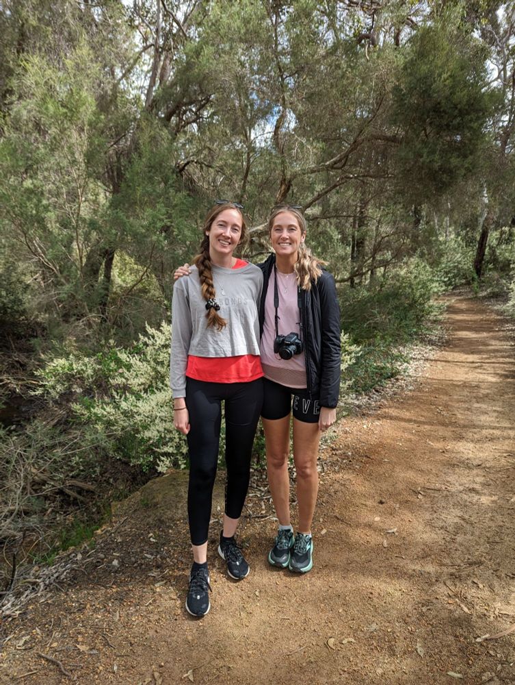 Adelle, wearing a grey sweater, pink top underneath and black leggings with runners and her twin sister wearing shorts, a pink
top, and a black jacket with a camera around her neck standing on a gravel path in the bush