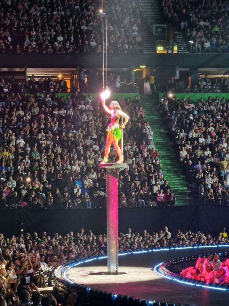 Katy perry standing on raised platform touching a lit heart hanging down.