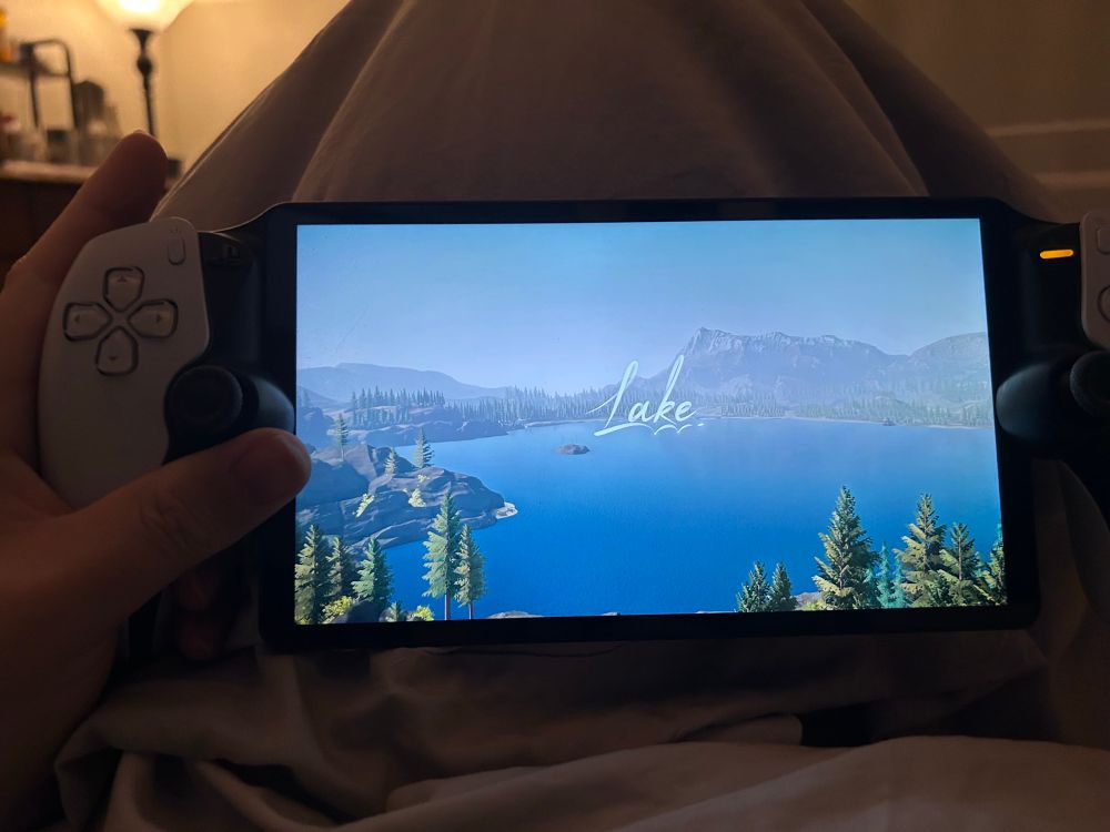 title screen for the game Lake displayed on a playstation portal handheld. the scene is a high wide shot overlooking a lake surrounded by evergreens and some mountains in the distance.