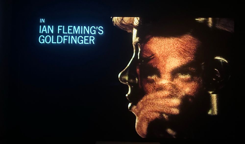 title screen that says IAN FLEMING'S GOLDFINGER. bond’s face is reflected over the face of a gold woman.
