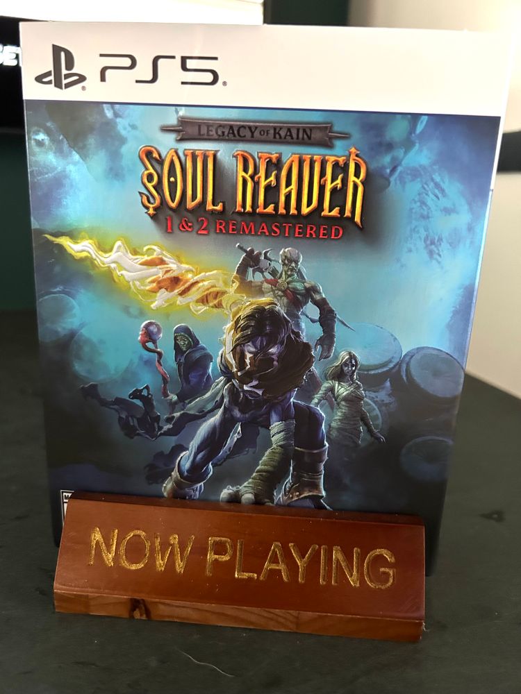 the PS5 case for soul reaver 1 and 2 remastered on a now playing stand. the cover art has raziel bent over with one arm on the ground while another arm is held out with the lighted up reaver wrapped around his arm. other characters are in the background.