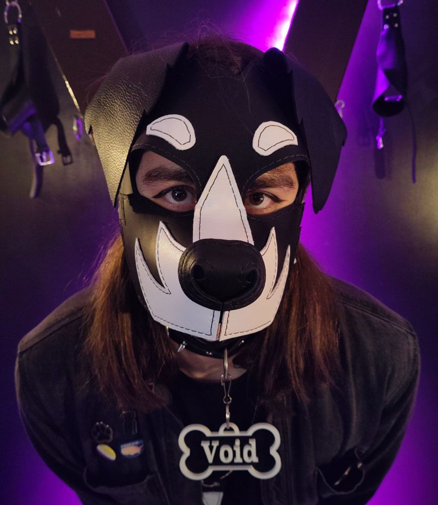 a person in black denim in front of a st. andrew's cross. they're wearing a leather puppy mask and a "Void" collar tag. they look directly at you, as if their pleading 