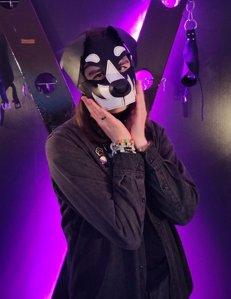a person in black denim in front of a st. andrew's cross. they're wearing a leather puppy mask holding their hands up to their face all innocently.