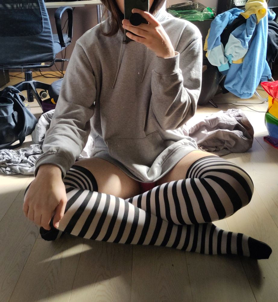 person taking a mirror selfie of them sitting on the floor wearing striped thigh highs, and a hoodie. they have their legs crossed
