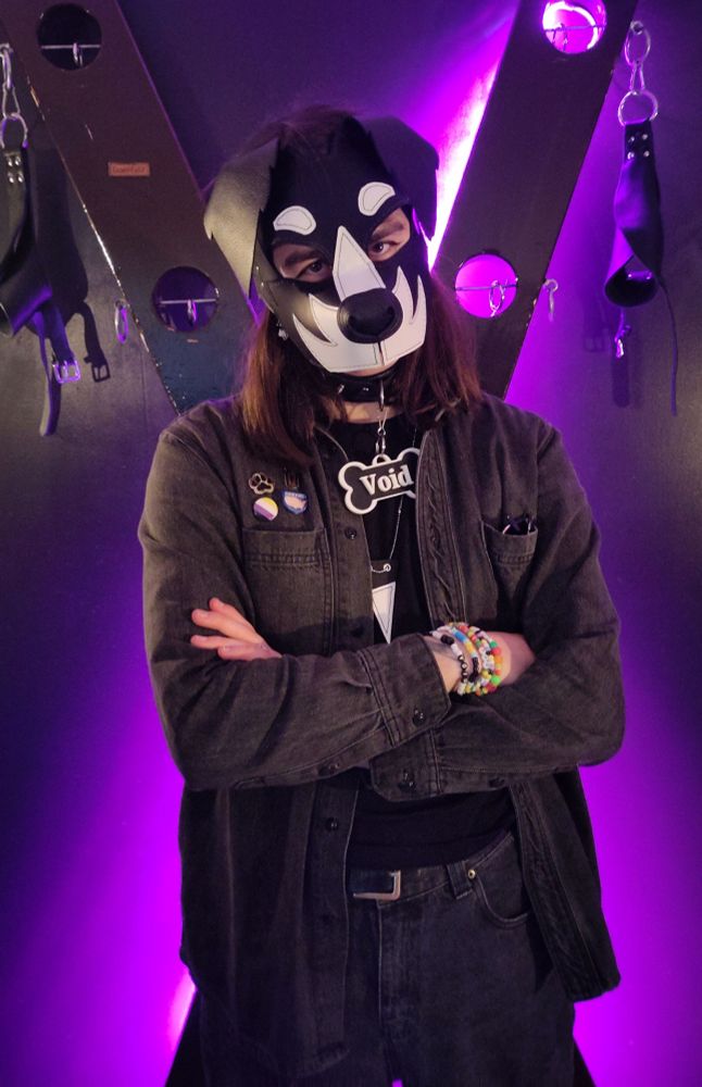a person in black denim in front of a st. andrew's cross. they're wearing a leather puppy mask with, as if they're playfully disappointed with you 