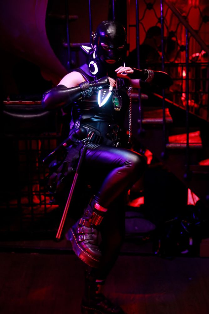 A person covered in latex and vinyl wearing a harness, LED headphones arround the neck. Holding by their hand an LED Kunai. 
They are slowly raising their foot in a combat position.