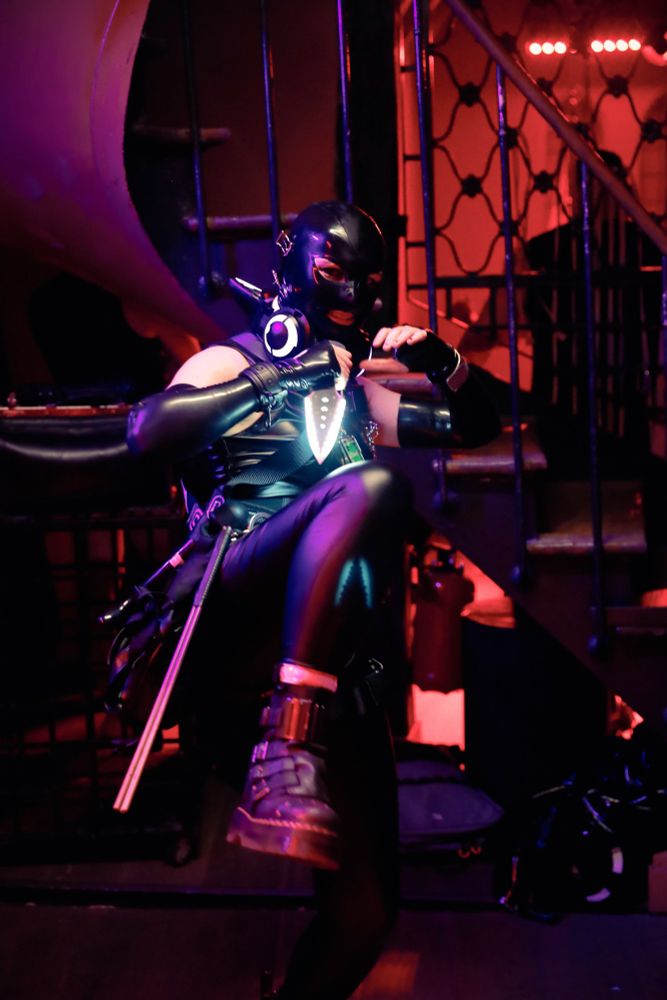 A person covered in latex and vinyl wearing a harness, LED headphones arround the neck. Holding by their hand an LED Kunai. 
They are slowly raising their foot in a combat position.