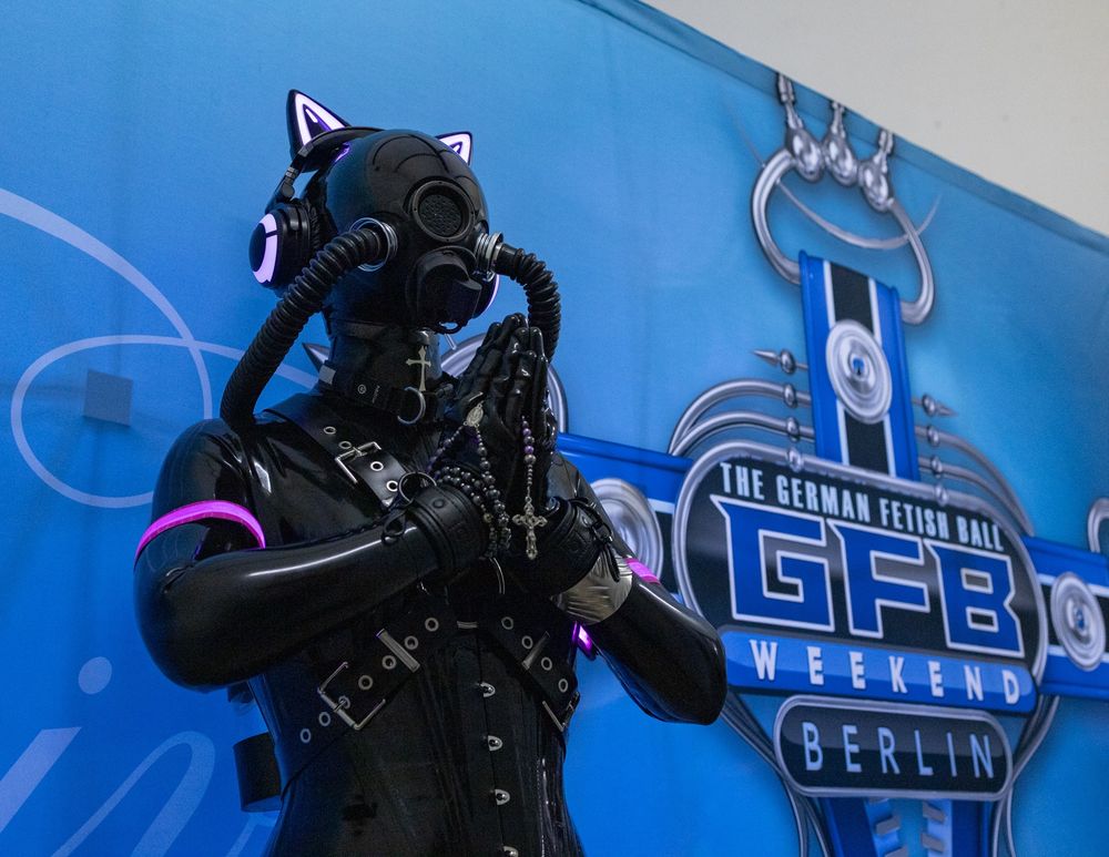 A latex drone wearing a heavy harness and two tubes on its gasmask. It is lifting its hands in prayer, holding a rosary.