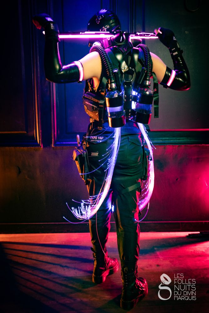The back of someone, they are holding a glowing purple word on their shoulder. They have heavy gear with tubes and LED filaments running on their back. The look is military inspired.