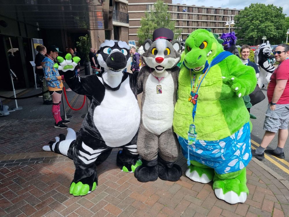 A large black and white plush alligator fursuit with green claws and blue eyes, a grey and white badger fursuit with yellow eyes, a red nose and a top hat and a large green crocodile fursuit with yellow eyes wearing blue and white shorts.