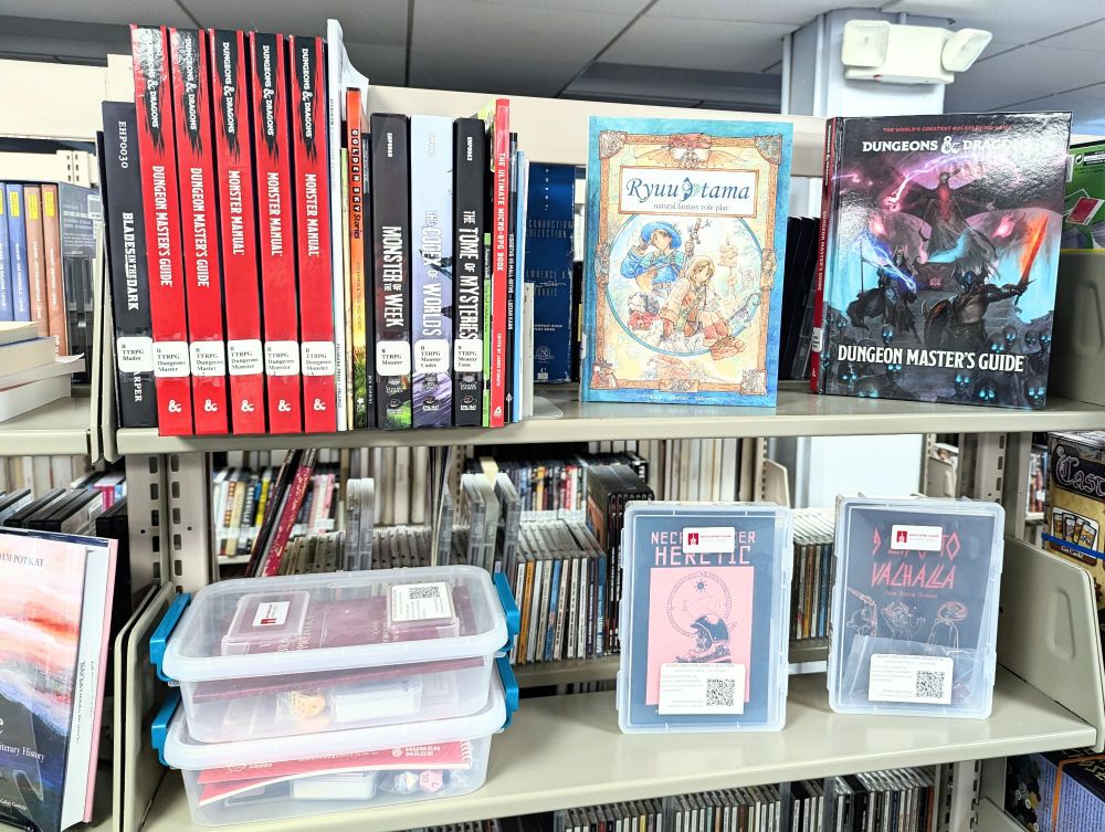 The top two shelves of a bookshelf, filled with TTRPGs. The top shelf contains several books. The second shelf holds small plastic totes that contain player kits for small, indie TTRPGs.
