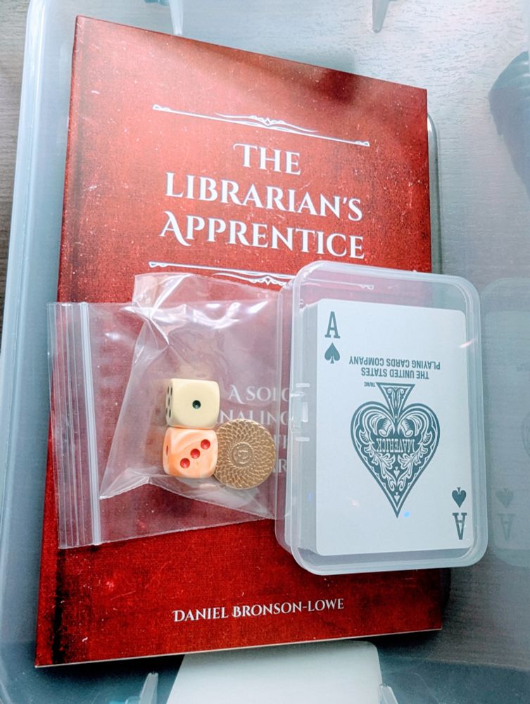 The game The Librarian's Apprentice in a plastic bin with cards, dice, and a coin
