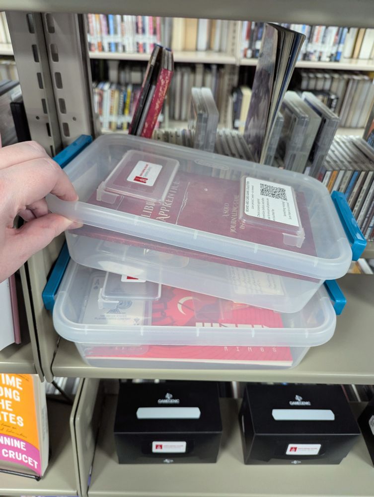 A game called The Librarian's Apprentice, in a plastic tote. Another tote underneath it contains a have called Moon Rings.