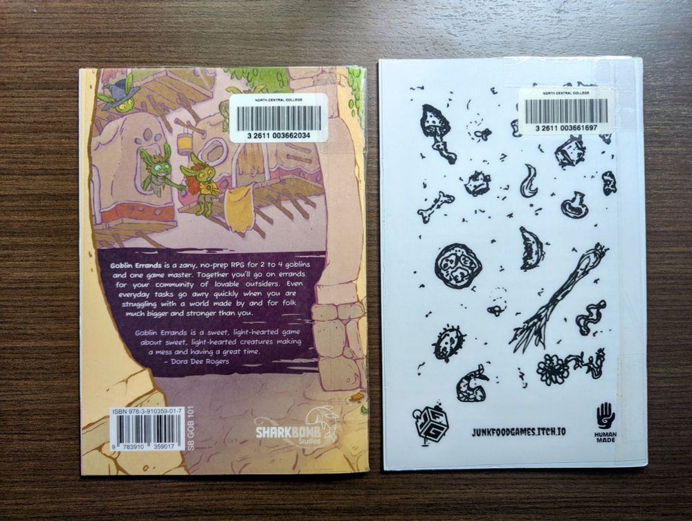 Two TTRPGs laying on a table, side by side, back cover up. Goblin Errands and Death Cap Saute.