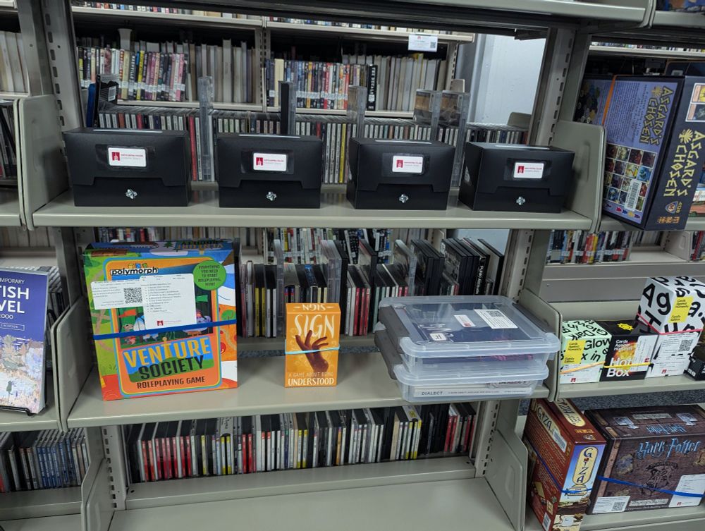 The bottom half of a bookshelf. The top pictured shelf contains player kits that hold tools that work across s wide array of games: a dice set, a coin, a character standee. The middle shelf contains TTRPGS that come in boxes and a couple plastic totes with player kits.