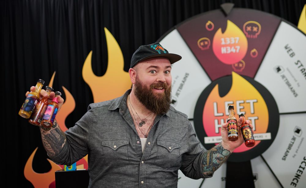 Jason standing in front of a game show where, holding five bottles of hot sauce. he has a slightly manic look in his eyes